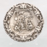 Prato de parede confeccionado em metal espessurado á prata. Formato circular, aba moldada em relevo por volutas, folhagens e motivos de acanto. Centro ornamentado por cena de interior em alto-relevo, representando quatro figuras em ambiente doméstico, com mobiliário, utensílios, tecidos e objetos suspensos ao fundo, em composição de caráter historicista inspirada em interiores europeus dos séculos XVI-XVII. Superfícies parcialmente brunidas, com contrastes entre áreas polidas e rebaixos escurecidos, acentuando a modelagem. Reverso reproduz em negativo a decoração obtida por estampagem, provido de argola para suspensão.Medidas: Diam.: 26 cm.