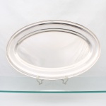 MERIDIONAL. Elegante travessa confeccionada em metal espessurado à prata, formato oval, centro ligeiramente rebaixado, aba largam, borda percorrida por contorno relevado. Marca da manufatura no verso. Medidas: Comp.: 67 x Larg.: 42,5 cm.