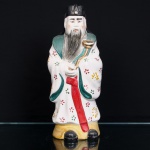 Escultura chinesa executada em porcelana moldada e esmaltada, representando Lu Xing, divindade associada à prosperidade, posição social e dignidade oficial na tradição chinesa, integrante do conjunto Fu Lu Shou. Figura masculina barbada, trajando veste cerimonial com mangas amplas, decorada com motivos florais policromados em vermelho, verde e amarelo, gola em verde, faixa frontal vermelha e toucado de oficial em preto. Segura nas mãos um ruyi, cetro de formato sinuoso, com acabamento dourado. Base em amarelo e calçados em preto. Marca caligráfica em vermelho na base. Medidas: Alt.: 29 x Comp.: 12 x Larg.: 9cm.