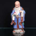 Escultura chinesa executada em porcelana moldada e esmaltada, representando Shou Xing, divindade taoista da longevidade, integrante do conjunto iconográfico Fu Lu Shou. Representado como ancião de fronte proeminente, crânio alongado, sobrancelhas e barba longas, trajando veste talar esmaltada em azul, com mangas amplas, reservas circulares em vermelho e ouro e debruns em vermelho e dourado. Sustenta na mão direita pêssego da imortalidade(shoutao), com folhas esmaltadas em verde, atributo associado à longevidade, e apoia a mão esquerda sobre cajado, outro elemento de sua iconografia. Decoração policromada sob e sobre o vidrado, em esmaltes azul, vermelho, verde, branco, preto, cinza e tons de carnação, com aplicação de dourado. Base moldada simulando formações rochosas, com esmaltação em cinza, preto, branco e vermelho. No funo da base, duas marcas com caracteres chineses, uma em baixo relevo e outra aplicada em vermelho, além de abertura circular de cocção. Medidas: Alt.: 25,5 x Comp.: 13 x Larg.: 9cm