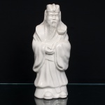 Escultura chinesa executada em porcelana, ao estilo Blanc de Chine, revestida por esmalte vítreo branco, representando Lu Xing, divindade associada à prosperidade, posição social, carreira e dignidade oficial, integrante da tríade Fu Lu Shou (), personificação das três aspirações de fortuna, prosperidade e longevidade. Na iconografia chinesa, Lu Xing é representado como um mandarim ou alto funcionário imperial, trajando vestes talares e toucado de oficial, atributos relacionados ao antigo sistema burocrático chinês e à obtenção de cargos e rendimentos. Figura masculina em pé, com barba longa, toucado e vestes de mangas amplas, modeladas em pregas. Segura diante do corpo um ruyi, cetro de formato sinuoso associado à autoridade, ao cumprimento dos desejos e à prosperidade. Base integrada à figura, com orifício de queima na parte inferior. Medidas: Alt.: 28,5 x Comp.: 12 x Larg.: 9cm