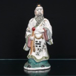 Escultura executada em porcelana chinesa representando Fu Xing, divindade associada à boa fortuna e integrante do conjunto Sanxing (Fu, Lu e Shou). Personagem masculino em posição ereta, com barba longa e toucado preto, trajando veste talar esmaltada em branco, decorada por ramagens estilizadas em verde e dourado, além de trigramas do Bagua delineados em preto. Segura bastão na mão direita e tecido drapeado sobre o braço esquerdo. Base moldada em tonalidade turquesa. Policromia em azul, rosa, vermelho, ocre, verde, preto e dourado. Marca em caracteres chineses na cor vermelha sob a base. Medidas: Alt.: 27,5 x Comp.: 13 x Larg.: 9cm
