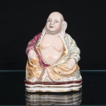 Escultura executada em porcelana chinesa representando Budai, monge da tradição Chan e personagem associado à abundância e contentamento, difundido na iconografia popular como o Buda Sorridente. Representado sentado, com cabeça calva, lóbulos auriculares alongados, torso parcialmente descoberto e vestes drapeadas. Manto com esmaltação em tons de rosa, verde-claro e ocre, ornamentado por motivos curvilíneos e florais, com realces e filetes dourados. Calças em tom ocre e base escalonada com faixa em vermelho e frisos dourados. Rosto, mãos, pés e torso em tonalidade marfim rosada, com detalhes fisionômicos delineados. Medidas: Alt.:15,5 x Comp.: 11,5 x Larg.: 10cm