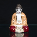 Escultura executada em porcelana esmaltada representando Buda sentado em postura meditativa, com mãos repousadas ao colo e vestes de panejamento modelado. Manto em tonalidade ocre, calças em vermelho-carmim e áreas em esmalte nacarado, complementadas por filetes e apontamentos dourados. Peça oca, com abertura de queima na base. Medidas: Alt.: 13,5 x Comp.: 9 x Larg.: 8cm