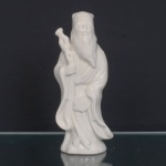 Escultura representando Shou Xing executada em porcelana branca esmaltada, moldada segundo a iconografia de Shou Xing, divindade estelar da longevidade e integrante da tríade Fu Lu Shou. Representado como ancião de barba longa, barrete e túnica de mangas amplas, portando cajado nodoso, atributo recorrente de sua representação. Corpo oco sobre base irregular com abertura circular de queima. Produção chinesa do século XX, segundo a tradição das porcelanas monocromáticas de Dehua, Fujian, difundidas no Ocidente sob a denominação 'Blanc de Chine'. Medidas: Alt.: 25,5 x Comp.: 10,5 x Larg.: 8cm