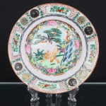 Prato executado em porcelana chinesa, decoração policromada e dourada executada em esmaltes sobre o vidrado, ao gosto da Família Rosa. Reserva central delimitada por cartela lobulada, contendo composição com tigre e ave de rapina, entre pinheiro, formações rochosas, flores e vegetação. Aba ornamentada por painéis alternados com frutos e motivos florais, intercalados por medalhões circulares contendo caracteres auspiciosos, além de frisos geométricos em vermelho-ferro e negro. Contorno realçado por filete alaranjado. No reverso, marca quadrangular em vermelho-ferro com quatro caracteres chineses(Huali Chang Zao).  Medida: Diam.: 21cm.