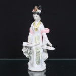 Figura chinesa de musicista em porcelana, século XX. Escultura moldada e esmaltada, com policromia em rosa, verde, amarelo, preto e douração, representando dama sentada sobre rochedo, envergando traje de mangas amplas, faixa envolvente e painéis com motivos fitomórficos. Penteado elevado guarnecido por flores aplicadas e ornato áureo. A personagem executa um guqin, cítara chinesa de sete cordas vinculada à tradição dos letrados(wenren). Composição complementada por roseira modelada em relevo, provida de caule, folhagem, botão e corola. Base irregular com abertura de queima e etiqueta oval inscrita MADE IN CHINA. Medidas: Alt.: 24,5 x Comp.: 13 x Larg.: 10,5 cm.