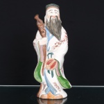 Escultura chinesa executada em porcelana moldada, esmaltada e policromada, representando Fu Xing, divindade estelar da felicidade e fortuna, integrante da tríade Fu Lu Shou. Personagem barbado, trajando túnica de mangas amplas com ramagens em vermelho-ferro, barrete e faixas em verde, azul e lilás, portando bastão nodoso e atributo circular suspenso por fitas. Corpo oco, base provida de abertura de queima e marca caligráfica estilizada em vermelho.Medidas: Alt.: 26 x Comp.: 10,5 x Larg.: 8 cm.