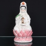Escultura executada em porcelana moldada, esmaltada e policromada, representando Guanyin, bodhisattva da compaixão no budismo chinês, sentada em padmasana sobre trono de lótus. Enverga manto de pregas descendentes e diadema com efígie de Amitabha(Amituofo) ao centro, atributo de sua iconografia. Mão direita elevada sustenta ramo de salgueiro, enquanto a esquerda ampara vaso kundika, recipiente associado à água purificadora. Peanha constituída por duas ordens de pétalas em rosa, delineadas e acentuadas a ouro. Corpo oco com abertura de queima na parte inferior.Medidas: Alt.: 29,5 x Comp.: 15 x Larg.: 11 cm.