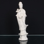 Escultura em porcelana branca moldada e esmaltada, representando Guanyin, bodhisattva da compaixão no budismo chinês, em posição ereta sobre pedestal com elementos florais modelados em relevo. Traja túnica de mangas amplas e panejamento longitudinal; cabelos reunidos em coque alto. Porta junto ao ombro flor de lótus, atributo recorrente em sua iconografia, enquanto a outra mão assume gesto ritual. Corpo oco com orifício de queima na base; marca impressa em verde, parcialmente apagada. Executada segundo a tradição das porcelanas brancas de Dehua(Fujian), conhecidas no Ocidente como Blanc de Chine. Medidas: Alt.: 29 x Comp.: 8,5 x Larg.: 8 cm