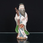 Escultura executada em porcelana moldada, esmaltada e policromada, representando Fu Xing, divindade da felicidade e fortuna, integrante da tríade Fu Lu Shou. Personagem barbado em posição ereta, trajando túnica de mangas amplas ornamentada por ramagens em vermelho-ferro, barrete, faixa azul e panejamentos em verde e lilás, ostenta bastão nodoso e atributo circular suspenso à cintura por fitas. Corpo oco, com abertura de queima na parte inferior e marca caligráfica estilizada em vermelho. Medidas: Alt.: 26 x Comp.: 11 x Larg.: 8 cm.