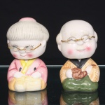 Duas(02) figuras em cerâmica moldada, esmaltada e policromada, representando casal de idosos japoneses sentado, com cabeças articuladas sobre mecanismo interno de mola, permitindo movimento oscilante. Ambos apresentam óculos circulares com lentes de vidro e armação metálica dourada. Figura masculina calva, com bigode, trajando quimono em amarelo reticulado, gola negra e parte inferior verde, segurando cachimbo tradicional japonês do tipo kiseru. Personagem feminina com cabelos brancos reunidos em coque, veste rosa sobreposta por gola amarela, mãos apoiadas sobre objeto modelado junto ao colo. Pintura em rosa, amarelo, verde, castanho, preto e dourado, com acentuação das faces em tonalidade rosada. Medidas: Alt.: 12 x Comp.: 8 x Larg.: 7cmMedidas: Alt.: 11,5 x Comp.: 7 x Larg.: 7cm