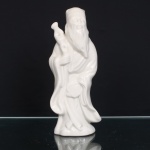 Escultura executada em porcelana branca moldada e esmaltada, representando Fu Xing, divindade da felicidade e fortuna, integrante da tríade Fu Lu Shou. Personagem barbado em posição ereta, trajando túnica de mangas amplas e barrete, portando bastão nodoso; à cintura, elemento circular suspenso por cordão. Corpo oco, base irregular com abertura de queima. Produção chinesa do século XX, segundo a tradição das porcelanas brancas de Dehua, Fujian, conhecidas no Ocidente como Blanc de Chine. Medidas: Alt.: 25,5 x Comp.: 11 x Larg.: 8cm
