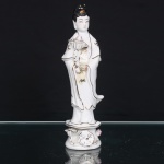 Figura em porcelana moldada e esmaltada, predominantemente branca, representando Guanyin em posição ereta, trajando túnica de mangas amplas com contornos e motivos fitomórficos realçados a ouro. Cabelos reunidos em coque, com ornamento dourado. Sustenta diante do torso flor de lótus sobre cálice foliáceo, enquanto a mão oposta apresenta gesto ritual. Assente sobre pedestal circular ornamentado em relevo com flores de lótus, folhagem e roda do Dharma(dharmachakra), símbolo budista alusivo aos ensinamentos de Buda. Corpo oco com abertura de queima na parte inferior. Medidas: Alt.: 21 x Comp.: 6 x Larg.: 5 cm