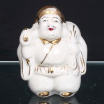 Escultura executada  em porcelana moldada e esmaltada, com douração e policromia pontual, representando Daikokuten, integrante dos Sete Deuses da Fortuna (Shichifukujin). Personagem de compleição corpulenta, trajando veste de mangas amplas e barrete, carregando às costas saco de riquezas, um de seus atributos iconográficos. Indumentária delineada a ouro, com faixa à cintura; cabelos em castanho, lábios em vermelho e feições demarcadas em preto. Corpo oco, com aberturas de queima na parte inferior. Medidas: Alt.: 11,5 x Comp.: 9 x Larg.: 8cm