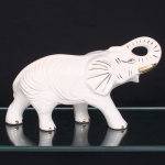Escultura em cerâmica moldada e esmaltada, esmaltação branca, representando elefante em marcha, com tromba elevada e recurvada. Modelagem anatômica definida por sulcos incisos no corpo, orelhas, membros e tromba; olhos, presas, unhas e demais elementos realçados a ouro. Corpo oco, com aberturas de queima sob as quatro patas. Medidas: Alt.: 18,5 x Comp.: 36 x Larg.: 11 cm.