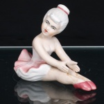 Escultura executada em porcelana moldada e policromada, representando jovem bailarina sentada em posição de alongamento, com pernas estendidas, braços projetados à frente e mãos apoiadas sobre os membros inferiores. Traja collant branco e tutu modelado em pregas, com gradações em rosa; sapatilhas na mesma tonalidade. Cabelos reunidos em coque, arrematado por faixa rosa. Fisionomia delineada, com olhos e sobrancelhas em preto e lábios em vermelho. Superfícies carnais com acabamento fosco, em contraste com áreas esmaltadas do traje. Base integrada à composição, formada pela própria saia. Medidas: Alt.: 8,5 x Comp.: 11 x Larg.: 6cm.