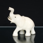 Escultura animalista talhada em pedra de tonalidade branco-leitosa, representando elefante em marcha, com tromba elevada, presas aplicadas e olhos constituídos por elementos esféricos negros incrustados. Anatomia definida por incisões que demarcam orelhas, tromba, pregas cutâneas e contornos corporais, superfície polida. Medidas: Alt.: 12,5 x Comp.: 13,5 x Larg.: 5,5cm.