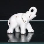 Escultura animalista em cerâmica branca esmaltada, representando elefante em marcha, com tromba elevada e recurvada, formando vão circular. Modelagem com orelhas em relevo, cauda aderente ao corpo e indicações anatômicas por sulcos incisos. Realces em dourado delineiam olhos, orelhas, unhas, boca e elementos do dorso. Corpo oco, com aberturas sob as patas decorrentes do processo de manufatura. Medidas: Alt.: 9 x Comp.: 10 x Larg.: 4,5cm.