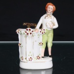 Paliteiro em porcelana. Peça escultórica em porcelana moldada e policromada, concebida como paliteiro, com figura masculina de pé junto a receptáculo arquitetônico, ornamentado por aplicações florais em relevo e filetes dourados. Personagem trajando calções verdes, camisa branca com laço ao pescoço e sapatos pretos, segurando pequeno livro junto ao peito. Base oval moldurada. Marca de manufatura em azul sob a base, fabricação nacional. Apresenta avaria visível nas fotos. Medidas: Alt.: 11 x Comp.: 7,5 x Larg.: 4cm.