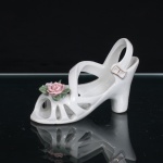 Miniatura de sapato feminino em porcelana esmaltada, modelada sob a forma de sandália de salto, com estrutura vazada composta por tiras arqueadas e contraforte elevado. Aplicação floral em relevo sobre a parte anterior, constituída por rosas em tonalidades rosa e lilás, acompanhadas de folhagem verde. Fivela lateral delineada a dourado. Medidas: Alt.: 7 x Comp.: 11 x Larg.: 4,5cm.