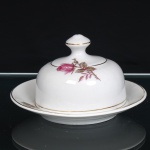 PORCELANA RAMI, Jundiaí - São Paulo(Fundada em 1930). Manteigueira em porcelana nacional, composta por prato circular, recipiente interno para acondicionamento e tampa campaniforme arrematada por pomo. Esmaltação branca, decoração floral com ramo de rosa em tonalidades rosa e verde, complementada por filetagem a ouro no contorno do prato, base e tampa. Marca de manufatura PORCELANA RAMI na base. Medidas: Alt.: 9 x Diam.: 15,5cm.