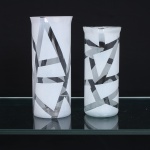 Dois(02) vasos em vidro incolor, de formato cilíndrico e alturas distintas, com paredes retas e bordas discretamente reviradas. Superfícies parcialmente fosqueadas, compostas por desenho geométrico abstrato formado por planos angulares, faixas sinuosas e reservas transparentes, em contraste com áreas de acabamento acetinado. Decoração de orientação modernista, executada por tratamento superficial do vidro. Medidas: Alt.: 20,5 x Diam.: 9cm; Alt.: 18,5 x Diam.: 8 cm.
