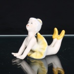 Figura de bailarina em porcelana, c.1950 -1970, representada em posição de alongamento, com o tronco elevado, braços estendidos à frente e pernas flexionadas para trás. Traja collant branco e saia de balé modelada em pregas, com esmaltação amarela, tonalidade repetida nas sapatilhas e no adorno do coque. Fisionomia delineada por pintura manual em preto e vermelho, com discretos matizes rosados na carnação. Peça moldada, oca, com orifício de respiro na base. Medidas: Alt.: 7,5 x Comp.: 10,5 x Larg.: 5,5cm.
