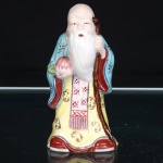 Escultura representando Shou Xing(Shoulao), divindade taoista da longevidade, executada em porcelana moldada e policromada. Representação de ancião de fronte elevada, longa barba branca e vestes talares em amarelo, azul, vermelho e rosa, ornamentadas por motivos florais e elementos estilizados. Porta cajado e pêssego da imortalidade (shoutao), atributos de sua iconografia e símbolos de longevidade na tradição chinesa. Base fechada com abertura de queima e numeração 253. Medidas: Alt.: 10,5 x Comp.: 5 x Larg.: 4,5cm.
