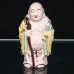 Escultura executada em porcelana esmaltada, representando Budai, monge associado na tradição chinesa à prosperidade, contentamento e abundância, posteriormente relacionado à figura do Buda Sorridente. Representado calvo, de ventre proeminente e semblante sorridente, trajando manto amarelo com gola azul-clara, ornamentado por motivos florais e elementos estilizados em policromia. Segura com ambas as mãos um cajado nodoso, disposto verticalmente junto ao corpo. Vestes em amarelo, rosa, azul, vermelho, preto e dourado. Base vazada. Medidas: Alt.: 10 x Comp.: 6 x Larg.: 4cm.