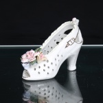 Escultura decorativa em porcelana, modelada sob a forma de sapato feminino de salto alto, com estrutura vazada em padrão reticulado geométrico. Parte frontal guarnecida por aplicação de flores e folhagens moldadas em relevo, nas tonalidades rosa, azul, creme e verde, com apontamentos em douradura. Laterais ornamentadas por motivos lineares dourados simulando elementos do calçado; abertura frontal e recortes junto ao calcanhar. Acabamento em esmaltação branca brilhante. Medidas: Alt.: 7 x Comp.: 9,5 x Larg.: 4,5cm.
