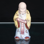 Escultura chinesa em porcelana esmaltada, representando Shou Xing(Shou Lao), divindade da longevidade e integrante do grupo dos Sanxing (Fu, Lu e Shou). Figurada como ancião de fronte proeminente e alongada, barba branca e semblante sereno, com as mãos recolhidas diante do corpo, encobertas pelas mangas. Traja túnica azul-clara e sobreveste amarela, ornamentadas por motivos florais estilizados, com detalhes em vermelho, rosa e azul. Cabelos brancos reunidos na nuca e presos por elemento verde. Base provida de abertura circular, decorrente da conformação oca empregada na manufatura da peça. Medidas: Alt.: 10 x Comp.: 5 x Larg.: 4cm.