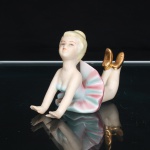 Escultura decorativa executada em porcelana, representando figura de bailarina, com o tronco elevado, braços estendidos à frente e pernas flexionadas para trás. Traja figurino de dança com corpete azul-claro e saia plissada, modelada em movimento, com gradações em rosa e azul. Cabelos loiros presos em coque; sapatilhas realçadas por douradura. Rosto com feições pintadas à mão em azul, alaranjado e castanho. Base inferior com pequeno orifício de respiro, próprio do processo de queima da peça oca, e numeração. Medidas: Alt.: 7,5 x Comp.: 10,5 x Larg.: 5,5cm.