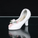 DYNASTY. Escultura representando sapato decorativo em porcelana esmaltada, modelado na forma de escarpim feminino de salto alto, com corpo branco e acabamento brilhante, gáspea ornamentada por aplicação floral em relevo, composta por rosa central e botões em tonalidades branca e rosa, acompanhados de folhagem verde; complementada por laço moldado em rosa. Interior côncavo, configurando pequeno recipiente decorativo. Etiqueta de manufatura DYNASTY aplicada na face inferior. Medidas: Alt.: 6,5 x Comp.: 10 x Larg.: 3,5cm.