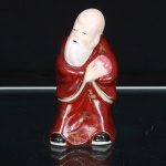 Escultura executada em porcelana esmaltada, representando Shou Lao (Shouxing), divindade taoista da longevidade e um dos três deuses estelares Fu Lu Shou. Apresenta crânio alongado e barba grande, segurando junto ao peito pêssego da imortalidade, atributo alusivo à longevidade. Traja manto em esmalte vermelho, decorado por motivos fitomórficos em douradura, com debruns em verde-claro; calçados em preto. Feições e elementos anatômicos delineados por pigmentação sobre o esmalte. Corpo moldado e oco, com abertura circular de queima na base.  Medidas: Alt.: x Comp.: x Larg.: