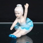 Escultura de bailarina em porcelana, modelada em posição sentada, com pernas estendidas, braço direito elevado e esquerdo projetado à frente. Figura feminina com cabelos presos em coque, feições pintadas à mão e traje de dança composto por corpete e saia de pregas onduladas, em esmaltes branco e azul-turquesa; sapatilhas na mesma tonalidade. Corpo parcialmente esmaltado, com acabamento nas áreas de carnação. Base integrada à composição, com orifício de queima e vestígios de numeração. Medidas: Alt.: 8 x Larg.: 10,5 x Comp.: 6cm.