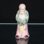 Escultura chinesa em porcelana, representando Shou Lao(Shouxing), divindade taoista da longevidade e personificação da estrela Shou, integrante do conjunto auspicioso Fu Lu Shou. Apresenta-se em pé, caracterizada pela fronte proeminente, sobrancelhas brancas, barba longa estriada e cabelos reunidos posteriormente. Traja túnica em esmalte rosa, com sobrevestimenta verde-clara, ambas ornamentadas por motivos florais e pequenos elementos fitomórficos; faixa frontal amarela e detalhes em vermelho, azul e douradura. Mãos recolhidas sob as mangas, segundo convenção iconográfica chinesa. Base oca, com abertura circular destinada à ventilação da peça durante a queima. Medidas: Alt.: 10,5 x Comp.: 4,5 x Larg.: 4,5cm.