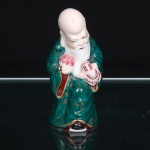 Escultura executada em porcelana moldada, esmaltada e policromada, representando Shou Lao, também denominado Shouxing, personificação da longevidade e integrante da tríade Fu Lu Shou. Figura reconhecível pela calota craniana alongada, cabelos recolhidos na nuca e barba que alcança o peito. Sustenta o pêssego da imortalidade, atributo associado à extensão da vida, e espécie de tartaruga estilizada, ornamentado por grafismos em vermelho e azul. Traja manto verde com motivos curvilíneos em douradura, forro das mangas em vermelho e calçados pretos. Modelagem oca, com abertura circular na base, decorrente do processo de conformação e queima. Medidas: Alt.: 10 x Comp.: 5 x Larg.: 4,5cm.
