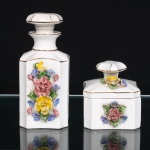 Conjunto de toucador em porcelana, composto por frasco de perfume e caixa para pó de arroz, ambos executados em cerâmica esmaltada, com corpos prismáticos de seção quadrangular e cantos chanfrados. Frasco de perfume com ombros facetados, gargalo estreitado e tampão encimado por pomo achatado; caixa com tampa ajustada, rematada por elemento de igual conformação. Decoração aplicada em relevo, constituída por rosas, botões, flores e folhagem modelados e policromados em rosa, amarelo, azul e verde. Contornos, gargalos, tampas e arestas realçados por filetes em douradura. Produção de meados do século XX, circa 19501960. Medidas: Alt.: 15 x Comp.: 6,5 x Larg.: 7cm. ; Alt.: 8,5 x Comp.: 7,5 x Larg.: 8cm.