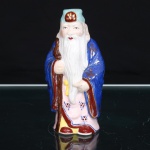 Escultura executada em porcelana esmaltada, representando Jurojin, divindade da tradição japonesa integrante dos Shichifukujin(Sete Deuses da Fortuna), associada à longevidade. Ancião de feições alongadas, barba longa e semblante sereno, portando cajado nodoso disposto verticalmente junto ao corpo.. Traja túnica em rosa sob manto azul, ornamentados por motivos florais e elementos estilizados em policromia, com apontamentos em douradura; barrete em azul-claro com reserva frontal em vermelho e ouro. Modelagem oca, com abertura de queima na base. Medidas: Alt.: 10,5 x Comp.: 5,5 x Larg.: 4cm.