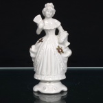 Figura feminina em porcelana esmaltada, de inspiração rococó, moldada em trajes de corte do século XVIII. Apresenta-se em pé, com penteado recolhido, corpete ajustado e saia de amplo volume, estruturada em pregas, panejamentos e festões; segura leque aberto na mão direita, mantendo a esquerda apoiada sobre o drapeado lateral. Superfície em esmaltação branca, com aplicações florais em douradura distribuídas pela indumentária. Assente sobre pedestal moldado de contorno ondulado, com ornamentação em relevo. Marca de manufatura ao fundo.  Medidas: Alt.: 14,5 x Comp.: 7 x Larg.: 5,5cm.