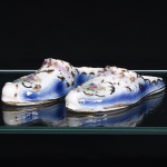 Par de sapatos ornamentais em porcelana, modelados à maneira de mules, com biqueiras fechadas, calcanhares abertos e bordas de contorno ondulado. Superfície esmaltada em branco, com gradações em azul e lavanda; ornamentação composta por reservas florais policromadas, circundadas por arabescos e elementos fitomórficos realçados a ouro. Região superior trabalhada em relevo com volutas e motivos de inspiração rocaille. Filetes dourados percorrem as extremidades e o contorno das solas. Interior provido de orifícios destinados à fixação à parede, apresentando em uma das peças a inscrição manuscrita Lar Feliz Apresentam craquelê, fissuras, desgastes e perdas pontuais de esmalte e douração, mais pronunciados em uma das biqueiras. Medidas: Alt.: 5,5 x Comp.: 20 x Larg.: 8cm.