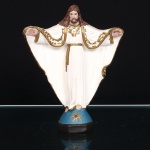 Escultura devocional de Cristo em estuque policromado. Figura de corpo inteiro representando Cristo, de cabelos longos e barba, trajando túnica branca de amplas mangas, com braços estendidos e palmas das mãos voltadas para a frente, assinaladas pelas chagas. Vestes enriquecidas por ornatos em dourado, com motivos fitomórficos e arabescados em relevo. Assenta sobre base escalonada, composta por seção superior azul com emblema radiante dourado e centro vermelho, sobre plinto inferior em tonalidade escura. Policromia em branco, castanho, azul, vermelho, rosa e dourado. Estado conforme fotografias. Medidas: Alt.: 19,5 x Comp.: 15,5 x Larg.: 5,5cm.