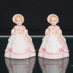 Duas(02) esculturas executadas em porcelana de biscuit esmaltada e policromada, representando damas em trajes românticos de época, segundo a tradição das figuras decorativas em estilo Crinolina/Vitoriano. Apresentam vestidos de saias volumosas, com corpete em rosa, saias em branco e rosa ornamentadas por pequenos pontos e laços em relevo, xales amarelos dispostos sobre os braços e amplas capotas(bonnets) presas por fitas sob o queixo. Mãos reunidas à frente, segurando pequeno elemento em forma de arco. Policromia em tons de rosa, amarelo, azul, castanho e vermelho, com realces dourados no corpete. Bases irregulares com orifício central. Sem marca de manufatura aparente. Medidas: Alt.: 8 x Comp.: 5,5 x Larg.: 5cm.