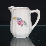 Leiteira em porcelana branca esmaltada, de corpo bojudo modelado com gomos verticais, bico vertedor pronunciado e alça lateral arqueada, com remate superior em relevo. Decoração policromada composta por ramalhetes florais, rosas e flores menores em tons de rosa, azul e amarelo, entre folhagens verdes. Filete dourado acompanha o contorno da borda e prolonga-se pela alça. Base circular reentrante. Sem marca de manufatura aparente. Medidas: Alt.: x Comp.: x Larg.: