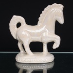Escultura de cavalo em faiança esmaltada. Figura equina modelada em posição de marcha, com uma das patas anteriores elevada, pescoço arqueado, crina e cauda definidas em relevo. Corpo recoberto por esmalte branco perolado de efeito nacarado, com reflexos iridescentes. Assenta sobre base oval moldurada por friso contínuo em relevo, de motivo entrelaçado. Apresenta pequenos desgastes e perdas pontuais no esmalte. Medidas: Alt.: 20,5 x Comp.: 19 x Larg.: 8,5 cm.