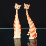 Duas(02) esculturas decorativas de gatos estilizados. Figuras felinas de corpo esguio e acentuadamente alongado, modeladas em posição sentada, com pescoços longilíneos, cabeças pequenas e caudas recurvadas junto às patas posteriores. Fisionomias estilizadas, com grandes olhos amendoados pintados, focinhos delineados e orelhas eretas. Superfície em tonalidade marfim, ornamentada por faixas irregulares em laranja, aplicadas em gradação aerografada, formando padrão alusivo à pelagem tigrada. Produção decorativa de estética Mid-Century Modern, circa 19501970. Sem marca de manufatura aparente. Medidas: Alt.: 15 x Comp.: 4 x Larg.: 3cm.
