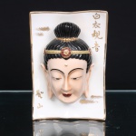 Placa devocional em porcelana chinesa representando Guanyin. Peça de formato retangular, com contorno suavemente ondulado e filetes dourados, apresentando ao centro máscara em alto-relevo de Guanyin(Avalokitevara), bodhisattva associado à compaixão no budismo do Leste Asiático. A representação exibe semblante contemplativo, olhos semicerrados, lóbulos auriculares alongados, urna assinalada na fronte e cabelos reunidos em coque elevado, ornado por diadema com motivos florais em vermelho e dourado. Fundo branco com inscrições caligráficas douradas: Báiyi Guanyin, correspondente a Guanyin de Vestes Brancas, e Shèngshan, Montanha Sagrada. Verso com marca de manufatura em azul e armação metálica destinada à fixação na parede. Medidas: Alt.: 17,5 x Comp.: 12 x Larg.: 6,5cm
