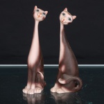 Par de esculturas decorativas representando gatos de formas estilizadas, executadas em material moldado, com corpos esguios e acentuadamente alongados, pescoços longilíneos, cabeças pequenas de feição antropomorfizada e caudas curvas junto às patas posteriores. Superfície em tonalidade marrom, com gradações em bege e rosado, faces policromadas, olhos amendoados e detalhes em preto e alaranjado. Modelagem característica da produção decorativa de inspiração Mid-Century Modern, difundida nas décadas de 19501960, marcada pela simplificação anatômica e pelo alongamento das proporções. Sem marca de manufatura aparente. Circa 19501960. Medidas: Alt.: 12 x Comp.: 5 x Larg.: 4cm.