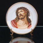 Porcelana Schmidt, Santa Catarina, Brasil (Fundada em 1945). Prato de parede em porcelana nacional, esmaltagem vítrea na cor branca, decorado com representação de Jesus Cristo coroado de espinhos, em composição de caráter devocional alusiva à Paixão, com cabeça voltada para o alto, cabelos longos, barba, marcas de sangue decorrentes da coroação e manto em tonalidade vermelha. Decoração policromada por transferência, com acabamento vitrificado. Aba delimitada por duplo filete dourado. No reverso, marca de fábrica sob esmalte: Porcelana Schmidt  Made in Brazil, encimada por coroa estilizada. Brasil, segunda metade do século XX. Medida: Diam.: 16,5cm.